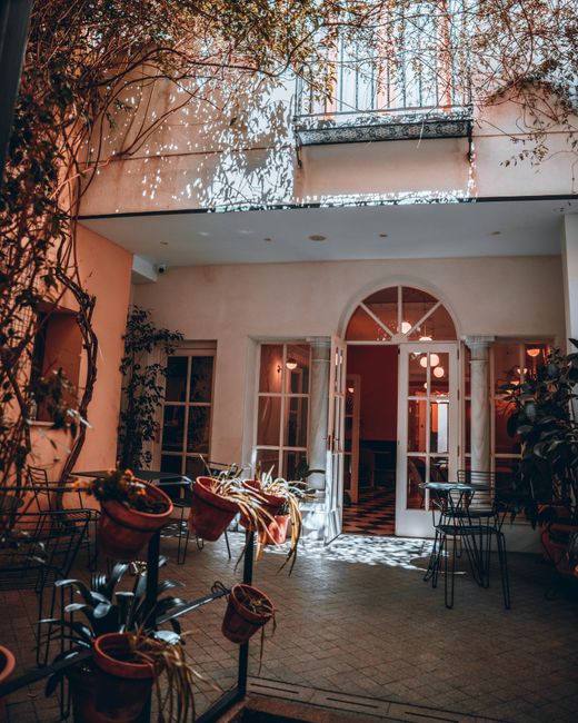 A cozy outdoor courtyard area featuring a mix of potted plants in various containers, with some pots directly resting on the brick-paved ground and others elevated on stands. The space is enclosed by