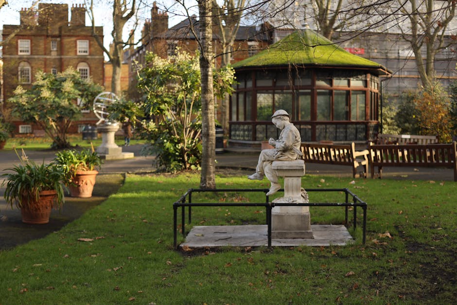 A serene garden scene showing a well-maintained lawn with lush, trimmed grass bordered by a variety of shaped shrubs and neatly pruned hedges. Tall, narrow evergreen trees are positioned towards the l
